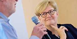 Ein Mann spricht in ein Mikrofon, während eine Frau mit kurzen blonden Haaren und Brille aufmerksam zuhört, ihr Kinn auf die Hand stützt und nachdenklich lächelt.