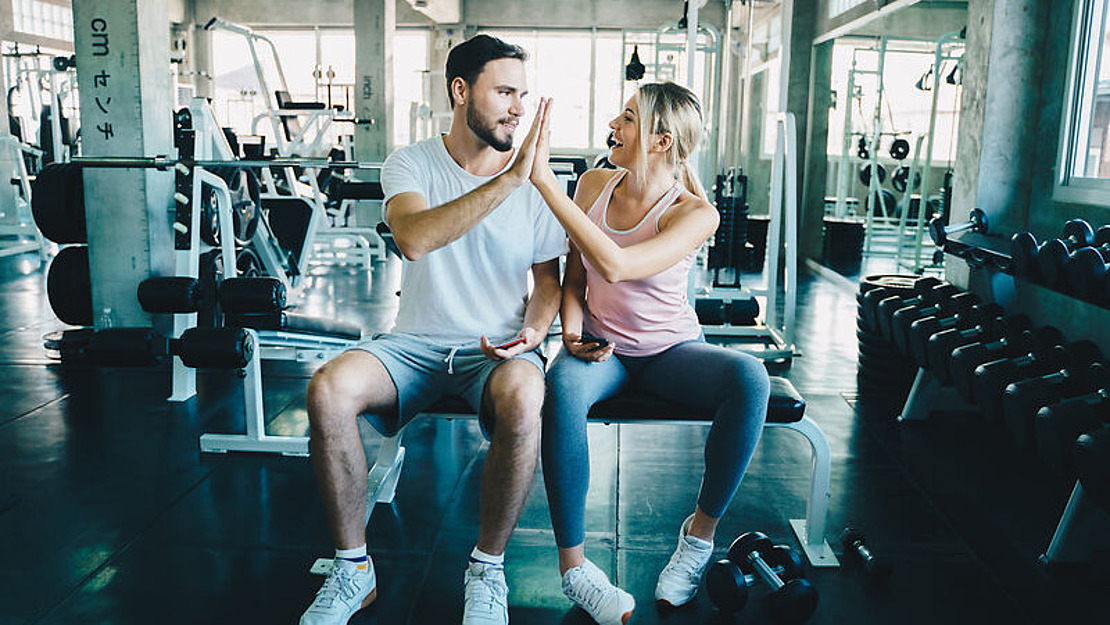 Ein Mann und eine Frau in Sportkleidung sitzen lächelnd auf einer Bank im Fitnessstudio und geben sich ein High Five. Im Hintergrund sind Trainingsgeräte und Gewichte zu sehen.