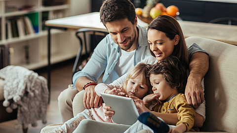 Eine vierköpfige Familie sitzt auf einem Sofa, lächelt und schaut gemeinsam auf ein Tablet. Die Eltern haben ihre beiden kleinen Kinder im Arm. Im Hintergrund steht ein Tisch mit Obst.