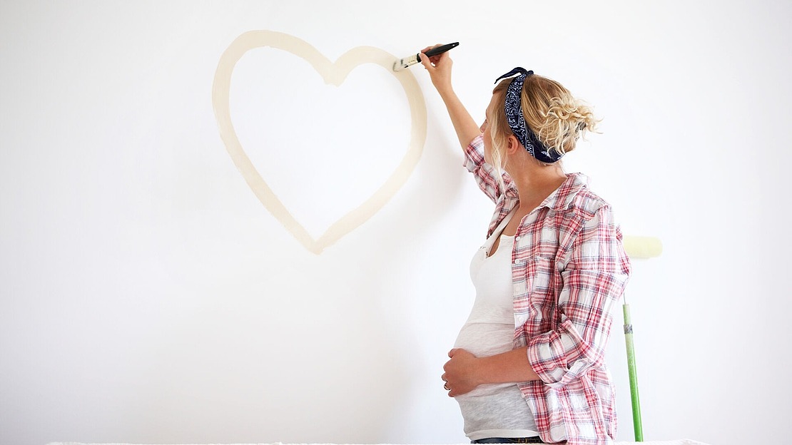 Eine schwangere Frau in einem karierten Hemd malt ein großes Herz an eine weiße Wand. Mit einer Hand streicht sie einen Pinsel, die andere stützt sie auf ihren Bauch. An der Wand lehnt eine Farbrolle.