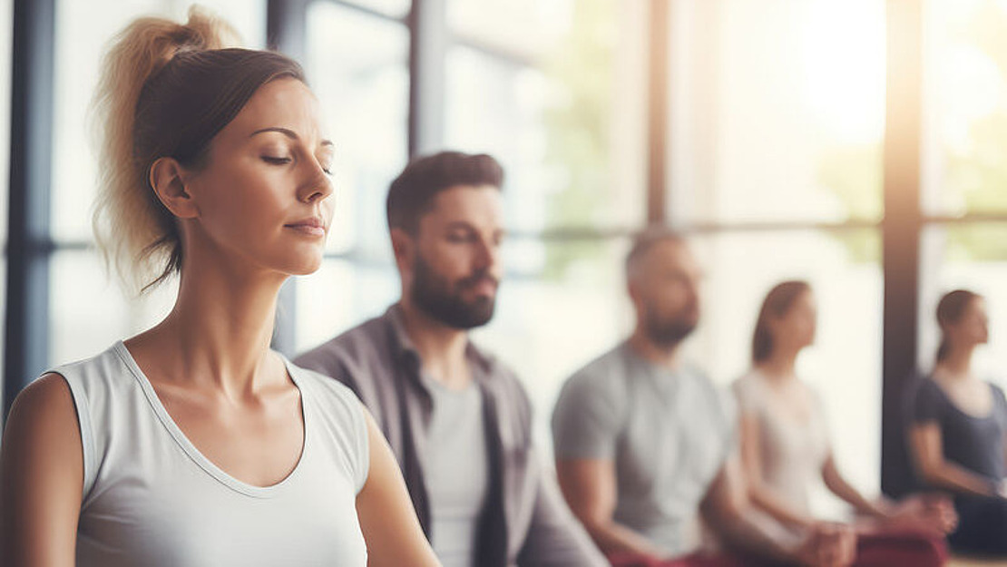 Eine Gruppe von Menschen sitzt im Schneidersitz auf Yogamatten in einem hellen Studio, die Augen geschlossen und meditierend. Der Fokus liegt auf einer Frau im Vordergrund, während andere Personen im Hintergrund verschwommen sind. Durch große Fenster strömt Sonnenlicht herein.