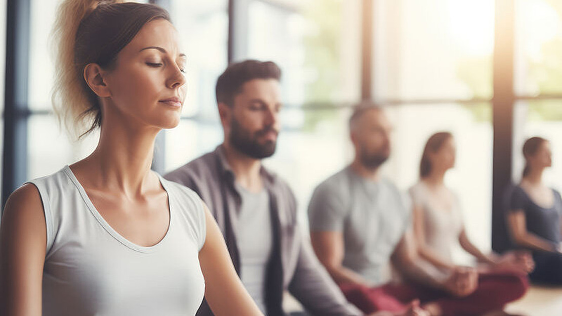 Eine Gruppe von Menschen sitzt im Schneidersitz auf Yogamatten in einem hellen Studio, die Augen geschlossen und meditierend. Der Fokus liegt auf einer Frau im Vordergrund, während andere Personen im Hintergrund verschwommen sind. Durch große Fenster strömt Sonnenlicht herein.