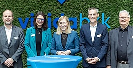 Fünf professionell gekleidete Personen stehen lächelnd hinter einem blauen Tisch, vor einer grünen Wand mit einem blauen Logo und teilweise sichtbarem Text im Hintergrund.