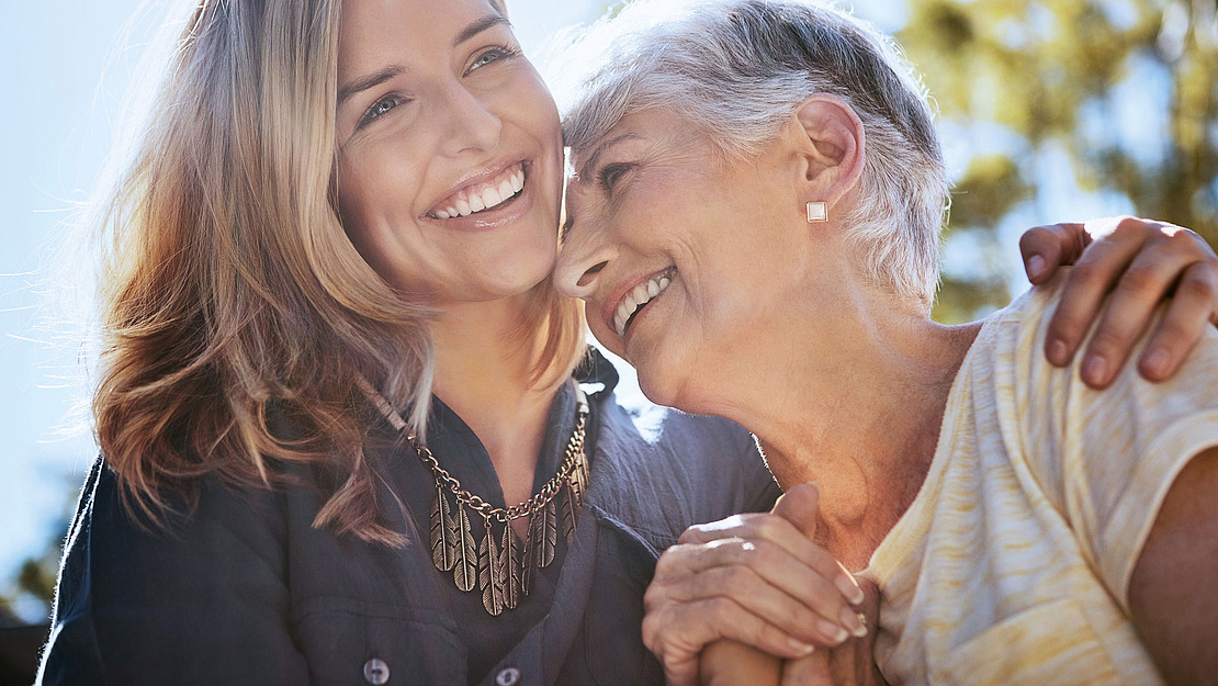Eine lächelnde junge Frau mit langen Haaren umarmt eine ältere Frau mit kurzen grauen Haaren. Sie sind draußen, das Sonnenlicht erzeugt einen warmen, leuchtenden Effekt. Beide tragen Halsketten und helle Hemden und teilen einen freudigen Moment miteinander.