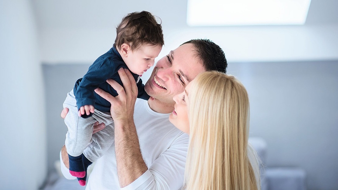Ein Mann und eine Frau lächeln fröhlich, während sie ein Baby im Arm halten. Der Mann hält das Baby an sein Gesicht, und die Frau blickt zu ihnen hinauf; alle wirken glücklich in einem hellen, sanft beleuchteten Raum.