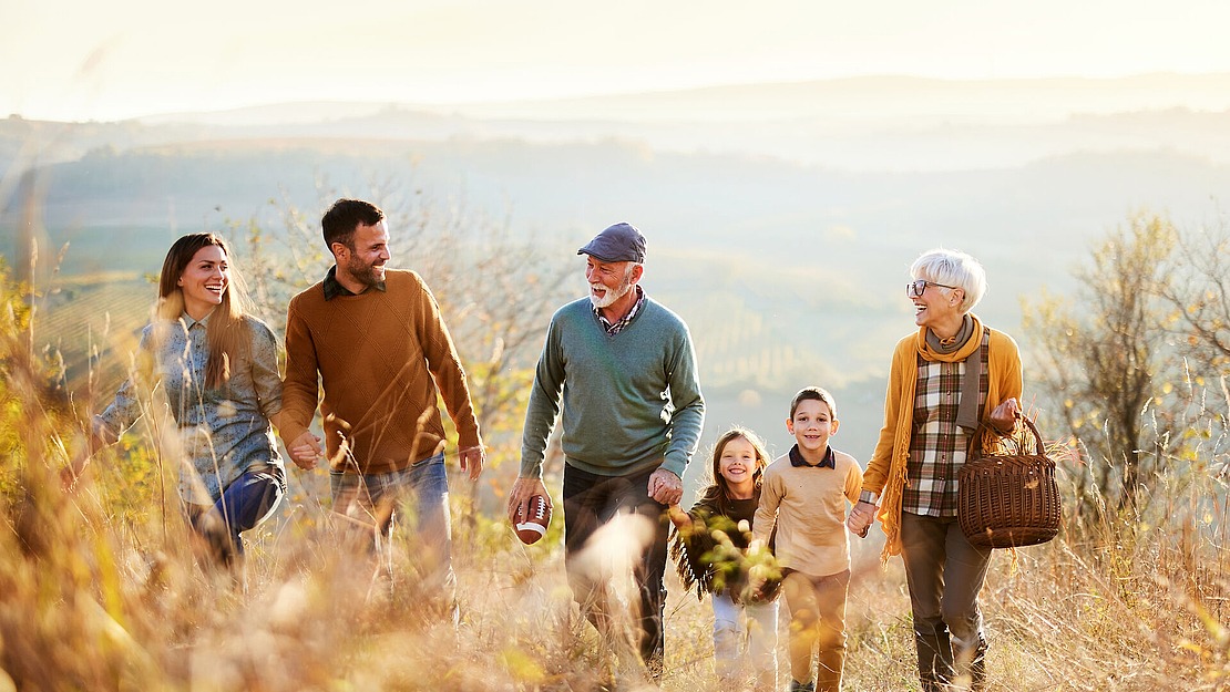 Eine sechsköpfige Familie, darunter zwei Kinder, geht gemeinsam durch ein malerisches, sonnenbeschienenes Feld. Sie tragen Herbstkleidung und lächeln, im Hintergrund sind sanfte Hügel zu sehen. Eine ältere Frau trägt einen Weidenkorb.