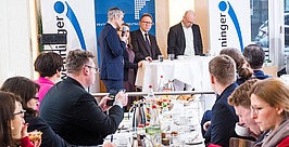 Eine Gruppe von Menschen nimmt an einer formellen Besprechung oder Podiumsdiskussion in einem hellen Raum teil. Vier Sprecher stehen vorne neben Bannern, während die Teilnehmer an Tischen mit Essen, Getränken und Notizbüchern sitzen.