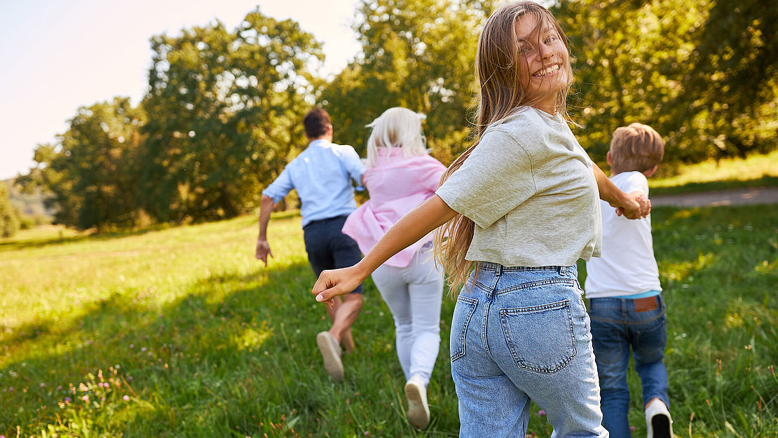 Ein lächelndes Mädchen in Freizeitkleidung dreht sich um, während es an einem sonnigen Tag Hand in Hand mit seiner Familie über eine Wiese läuft, umgeben von Bäumen und Grün.