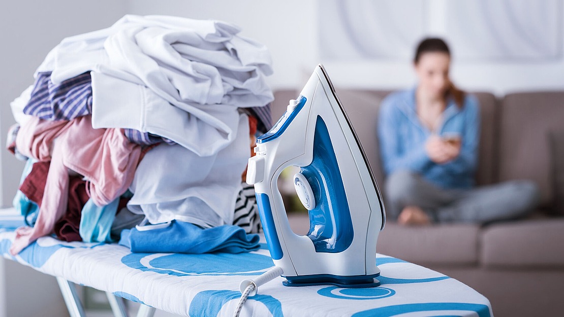 Ein Stapel Wäsche liegt auf einem Bügelbrett, im Vordergrund ein Dampfbügeleisen. Im Hintergrund sitzt eine Person auf einem Sofa und telefoniert. Die Szene lässt vermuten, dass gerade Wäsche gewaschen wird.