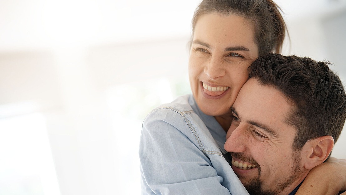 Eine lächelnde Frau umarmt einen lächelnden Mann in einem Haus. Beide wirken glücklich und blicken leicht aus der Kamera. Weiches natürliches Licht füllt den Hintergrund.
