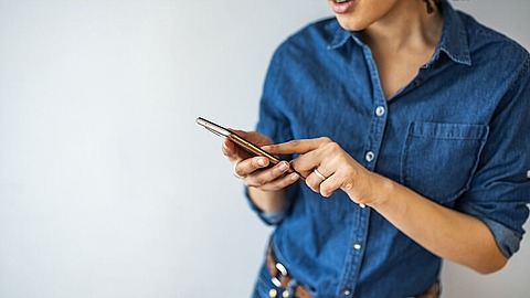 Eine Person, die ein blaues Jeanshemd und einen braunen Gürtel trägt, hält ein Smartphone in der Hand und tippt auf den Bildschirm, während sie vor einem schlichten hellen Hintergrund steht. Nur der Oberkörper und das untere Gesicht sind sichtbar.