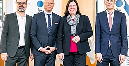 Vier professionell gekleidete Personen, drei Männer und eine Frau, stehen nebeneinander im Innenbereich und lächeln in die Kamera, im Hintergrund sind Banner und Fenster zu sehen.