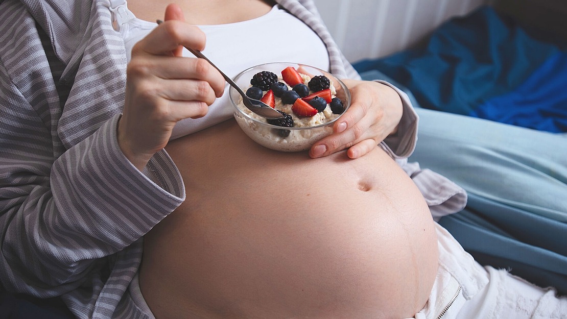 Eine schwangere Person liegt im Bett. Sie trägt einen grau gestreiften Morgenmantel und hält eine Schüssel Haferbrei mit Erdbeeren, Blaubeeren und Brombeeren in der Hand. Eine Hand hält einen Löffel über der Schüssel.