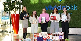 Fünf Frauen stehen drinnen vor einer grünen Wand mit dem Logo von vividabkk und halten verpackte Geschenke in den Händen. Vor ihnen auf dem Boden stapeln sich viele bunt verpackte Geschenke.