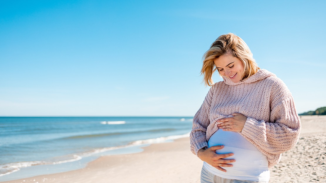 Eine schwangere Frau in einem leichten Pullover steht an einem Sandstrand, wiegt sanft ihren Bauch und lächelt, im Hintergrund das Meer und der blaue Himmel.