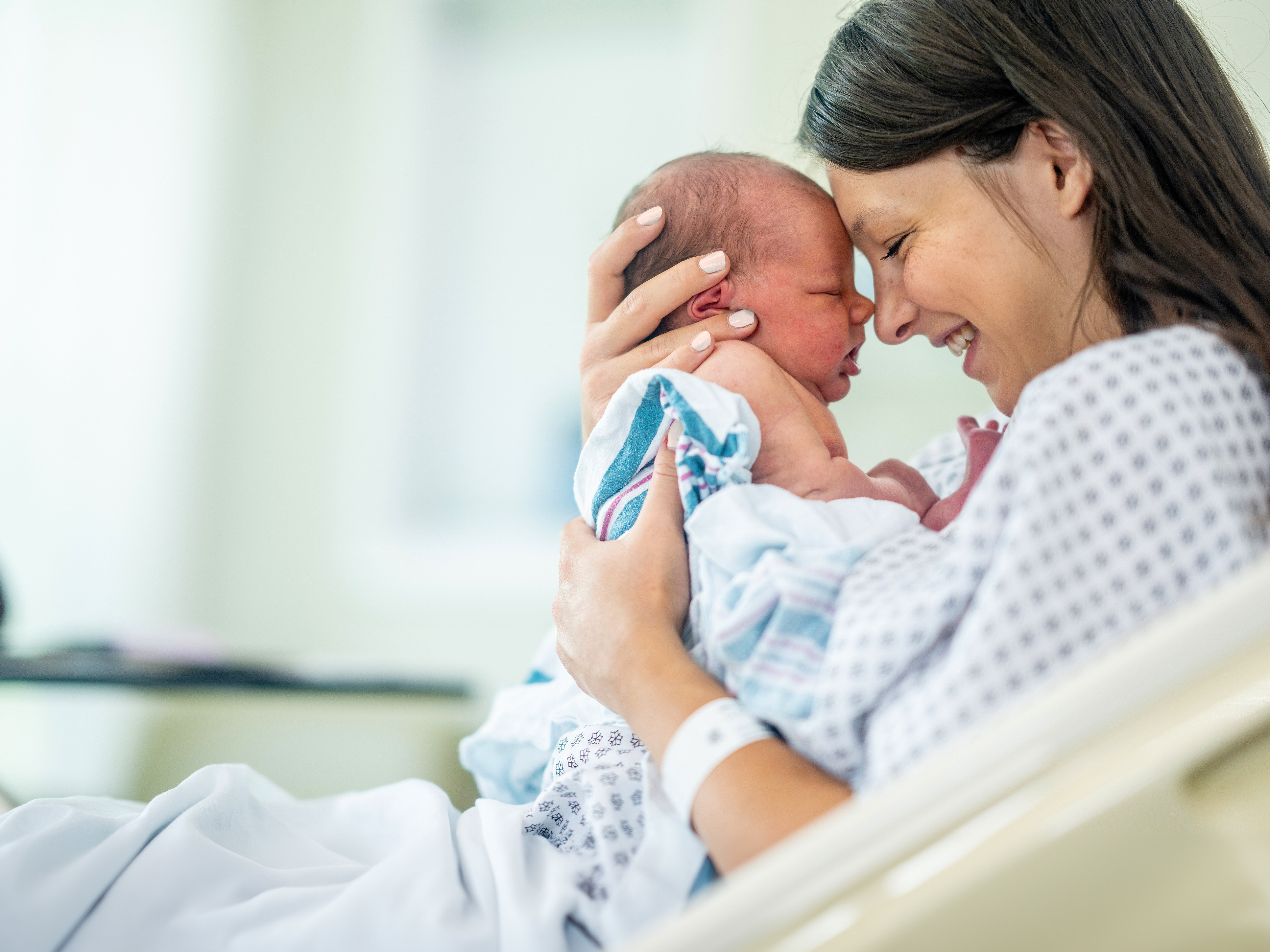 Eine lächelnde Frau im Krankenhaushemd hält ein in eine Decke gewickeltes Neugeborenes und berührt in einem hellen, sanft beleuchteten Krankenhauszimmer sanft die Stirn des Säuglings.