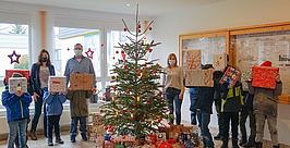 Eine Gruppe Kinder steht mit eingepackten Geschenken um einen geschmückten Weihnachtsbaum im Haus. Hinter ihnen stehen drei Erwachsene, alle mit Mund-Nasen-Schutz. Weitere Geschenke liegen unter dem Baum, und festliche Dekorationen sind zu sehen.