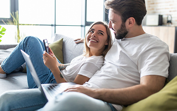 Eine Frau und ein Mann sitzen zusammen auf einem Sofa. Die Frau, die ein Telefon hält, lächelt, während sie zu dem Mann aufblickt. Der Mann, der einen Laptop hält, blickt sie an. Beide tragen weiße Hemden und Jeans. Im Hintergrund sind ein großes Fenster und eine Pflanze zu sehen.