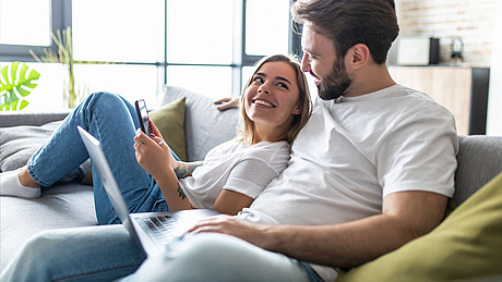 Eine Frau und ein Mann sitzen zusammen auf einem Sofa. Die Frau, die ein Telefon hält, lächelt, während sie zu dem Mann aufblickt. Der Mann, der einen Laptop hält, blickt sie an. Beide tragen weiße Hemden und Jeans. Im Hintergrund sind ein großes Fenster und eine Pflanze zu sehen.