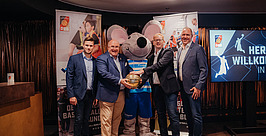 Vier Männer in Anzügen und ein großes Mausmaskottchen stehen im Innenbereich zusammen und berühren einen goldenen Basketball. Im Hintergrund sind Banner und ein Bildschirm mit Basketball-Grafiken zu sehen.