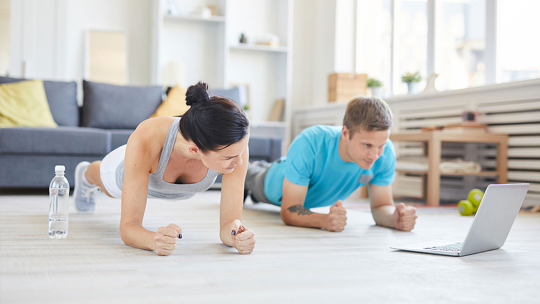 Eine Frau und ein Mann machen zu Hause Unterarmstützübungen auf dem Boden, vor einem Laptop-Bildschirm. Eine Wasserflasche steht daneben. Der Raum ist hell und modern, im Hintergrund ein Sofa und Regale.