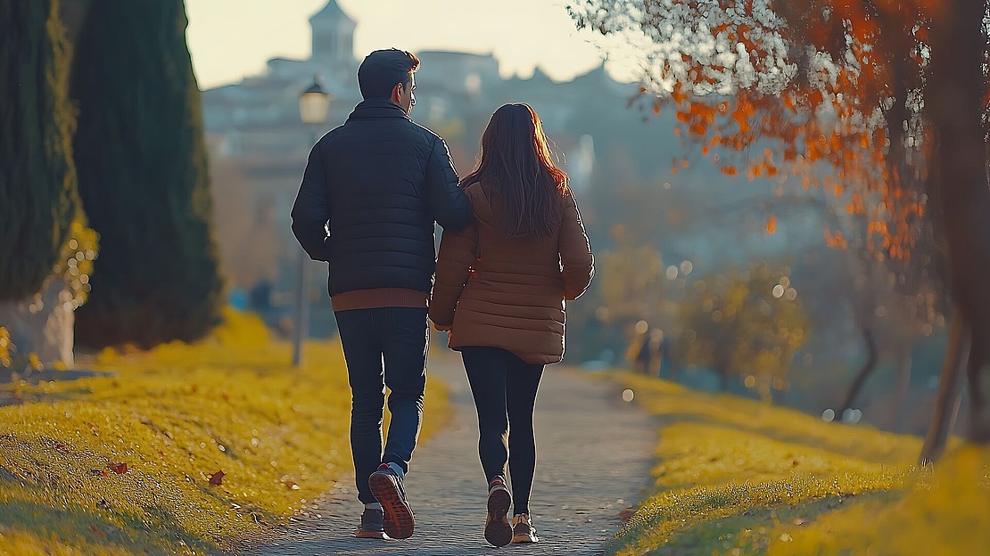Ein Paar in Jacken spaziert Hand in Hand einen von Bäumen gesäumten Weg in einem Park im Herbst entlang, mit gefallenen Blättern auf dem Boden und einer sichtbaren Stadt im Hintergrund.