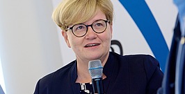 Eine Frau mit kurzen blonden Haaren und Brille spricht in ein Mikrofon. Sie trägt einen dunklen Blazer über einem gemusterten Oberteil. Hinter ihr ist eine verschwommene Figur vor einem blau-weißen Hintergrund zu sehen.