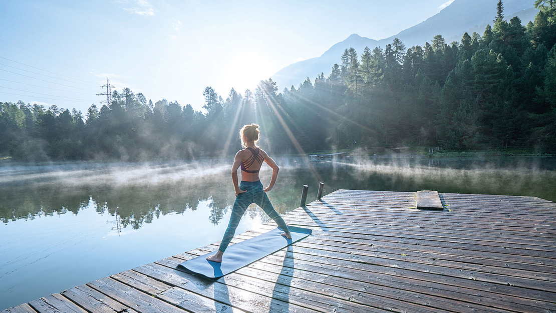 Eine Person steht in einer Yoga-Pose auf einer Matte auf einem Holzsteg an einem ruhigen See, umgeben von Bäumen und Bergen, während am frühen Morgen Sonnenlicht durch den Nebel strömt.