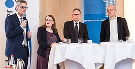 Vier Personen in Businesskleidung stehen an zwei runden Tischen und sprechen und hören während einer formellen Veranstaltung zu. Ein Mann hält ein Mikrofon. Auf den Tischen stehen Getränke, im Hintergrund ist ein Banner der „Deutschen Welle“ zu sehen.