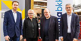 Vier Personen in Geschäftskleidung stehen vor zwei Bannern mit Logos und posieren für ein Gruppenfoto bei einer Indoor-Veranstaltung. Im Hintergrund sind Weinflaschen zu sehen.
