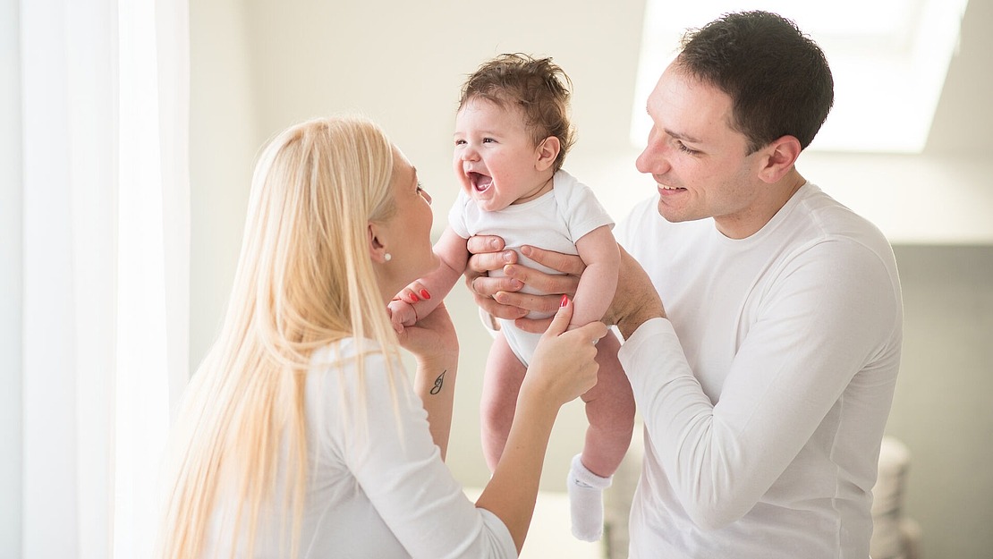 Ein lächelndes Baby wird von einem Mann gehalten, während eine Frau dem Baby gegenübersteht, alle tragen weiße Kleidung. Das Baby sieht glücklich aus und alle scheinen in einem hellen, sonnenbeschienenen Raum fröhlich zu sein.