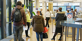 In einem modernen Gebäude mit Glaswänden und Holzboden stehen Menschen Schlange. Einige tragen Jacken und Taschen. Im Hintergrund sind Schilder, Tische und Mitarbeiter zu sehen.