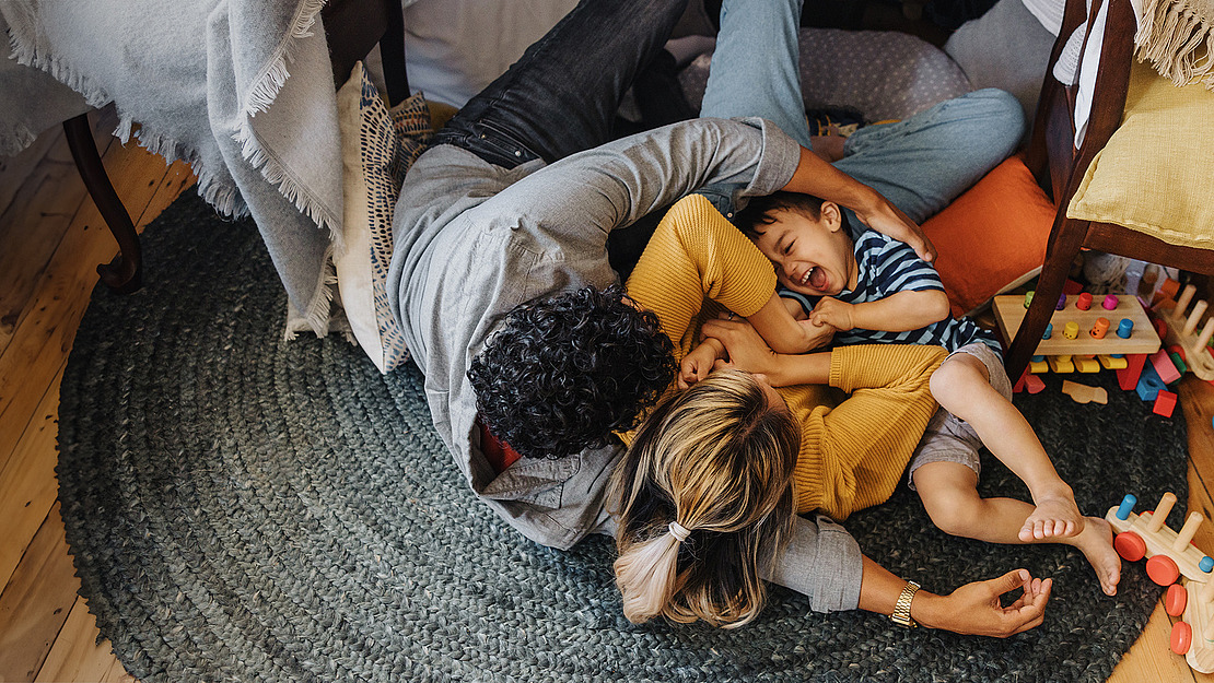 Eine Familie spielt zusammen auf einem runden grünen Teppich, lacht und kuschelt auf dem Boden zwischen verstreuten Holzspielzeugen und bunten Kissen in einem gemütlichen, sonnendurchfluteten Raum.