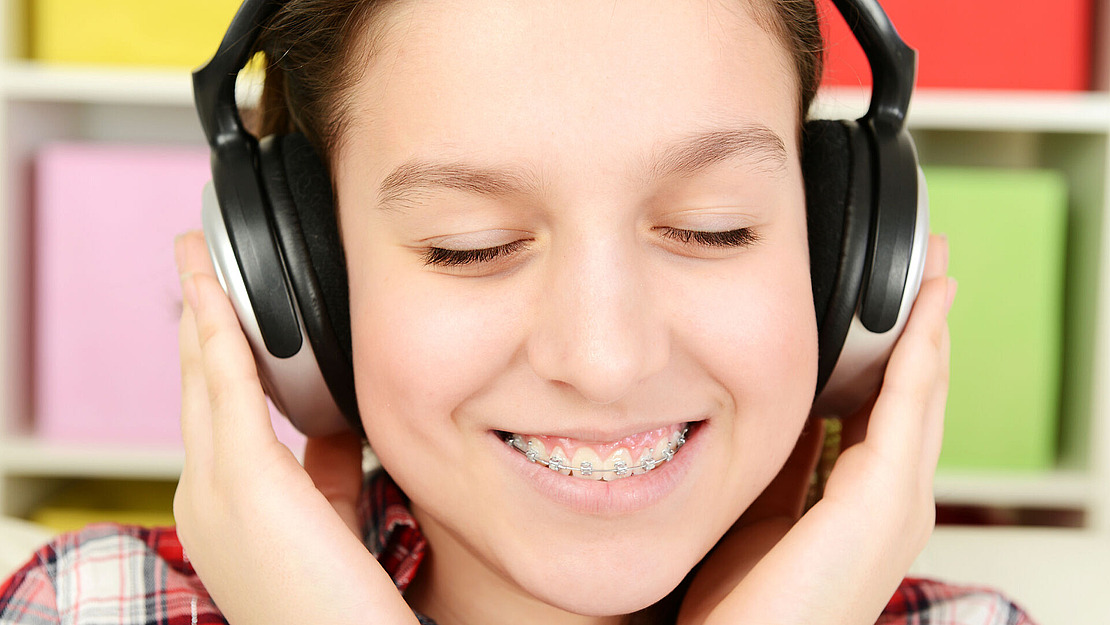 Eine Person mit Zahnspange lächelt mit geschlossenen Augen und hört Musik über große Kopfhörer. Sie hält beide Seiten der Zahnspange fest. Im Hintergrund sind bunte, verschwommene rechteckige Formen zu sehen.