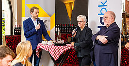 Ein Mann in einem blauen Blazer steht neben einem rotkarierten Tisch und hält ein Mikrofon in der Hand. Eine Frau und ein älterer Mann halten ebenfalls Mikrofone in der Hand und sprechen in einem Restaurant zu einem Publikum. Im Hintergrund sind Transparente zu sehen.