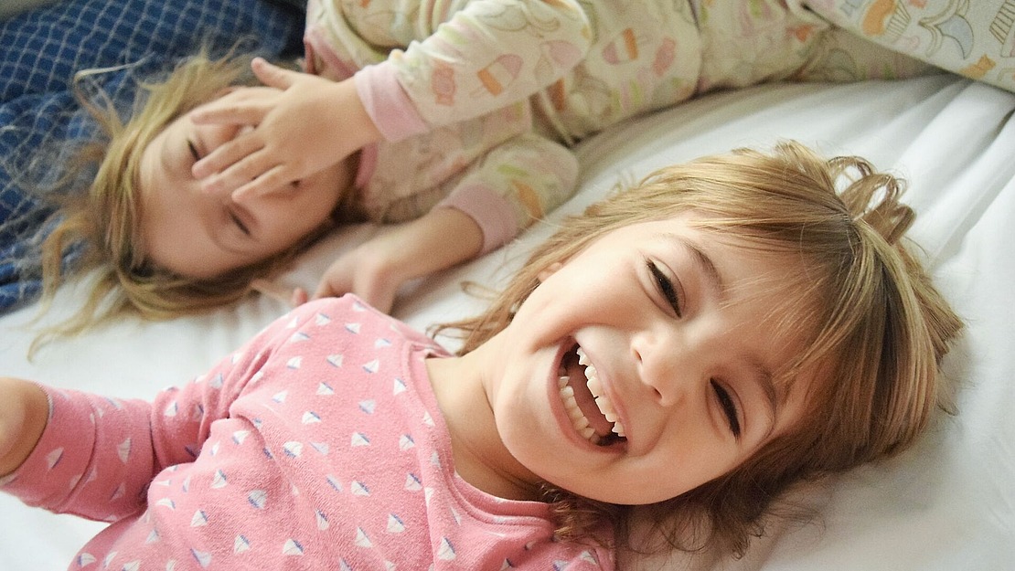 Zwei kleine Kinder in Pyjamas liegen auf einem Bett. Ein Kind in einem rosa Schlafanzug lächelt und lacht in die Kamera, während das andere Kind in einem gemusterten Schlafanzug ein Auge mit einer Hand bedeckt und entspannt aussieht.