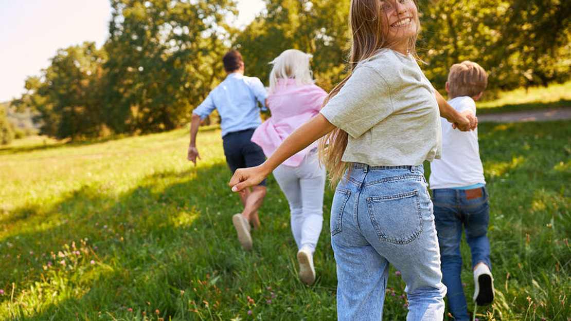 Eine vierköpfige Familie läuft an einem sonnigen Tag fröhlich über eine Wiese. Die Frau im Vordergrund lächelt in die Kamera. Bäume säumen den Hintergrund, und die Szene ist von leuchtendem Grün und warmem Licht erfüllt.