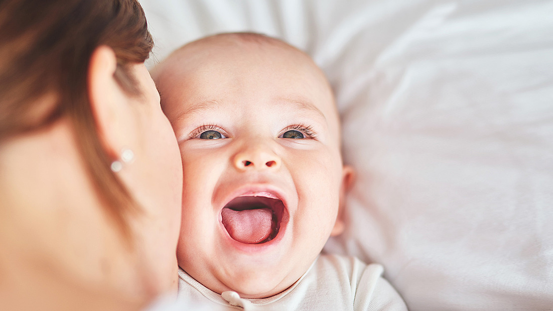 Ein lachendes Baby liegt auf einem weißen Laken, eine Person lehnt sich dicht an seine Wange. Die Augen des Babys leuchten und sein Mund ist vor Freude geöffnet. Die Szene vermittelt ein Gefühl von Wärme und Glück.
