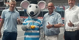 Drei Männer stehen auf einem Basketballplatz, lächeln und zeigen die Daumen nach oben neben einer Person in einem großen Maus-Maskottchen-Kostüm, die ein gestreiftes Hemd trägt und eine Medaille hält.