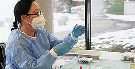 Eine medizinische Fachkraft mit Maske, Handschuhen und Kittel bereitet an einem Tisch mit medizinischem Material eine Spritze vor. Durch das Fenster dahinter ist eine verschneite Landschaft zu sehen.