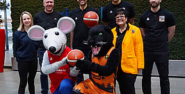Sieben Personen posieren im Innenbereich, darunter zwei in Maskottchenkostümen – einer grauen Maus und einem schwarzen Panther –, die beide Basketbälle in der Hand halten. Die anderen tragen Freizeit- oder Sportkleidung und stehen lächelnd vor einer grünen Wand.