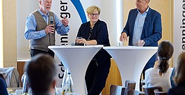 Drei Personen in Geschäftskleidung stehen während einer Veranstaltung an hohen runden Tischen und sprechen. Im Vordergrund sitzen Teilnehmer. Im Hintergrund sind zwei Banner mit blauen Logos zu sehen.