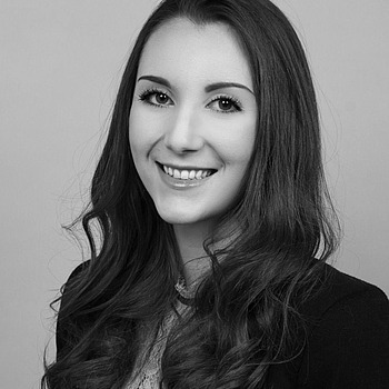 Schwarz-weißes Porträt einer jungen Frau mit langem, gewelltem Haar, lächelnd, in einem Spitzenoberteil und einem Blazer, vor einem schlichten Hintergrund.