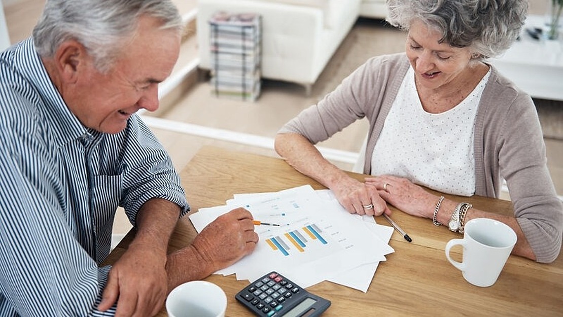 Ein älterer Mann und eine ältere Frau sitzen an einem Holztisch und prüfen Diagramme auf Papier. Auf dem Tisch stehen ein Taschenrechner und zwei Kaffeetassen. Sie scheinen in einem hell erleuchteten Raum Finanzdokumente zu besprechen.