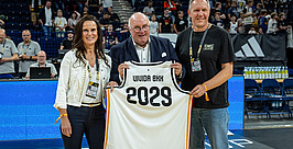 Drei Personen stehen auf einem Basketballfeld und halten ein Trikot mit der Aufschrift „2029“ und „Ulm/Old BKH“ in den Händen. Im Hintergrund sind Zuschauer und Veranstaltungsmitarbeiter zu sehen.