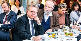 Eine Gruppe professionell gekleideter Personen sitzt während einer Indoor-Veranstaltung an Tischen und hört aufmerksam zu. Im Vordergrund stehen zwei Männer in Anzügen mit Getränken, Zeitungen und Essen auf dem weiß gedeckten Tisch.