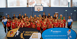 Eine große Gruppe von Kindern und Erwachsenen in rot-schwarzer Sportkleidung posiert in einer Turnhalle mit Basketballkorb und hält zwei Banner des Deutschen Basketball Bundes und von vividabkk hoch. Im Hintergrund sind Fenster und Turngeräte zu sehen.