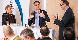 Drei Personen sprechen in einer Podiumsdiskussion vor Publikum. Ein Mann gestikuliert mit dem Daumen nach oben und hält ein Mikrofon in der Hand. Eine Frau und ein weiterer Mann halten ebenfalls Mikrofone in der Hand und diskutieren.