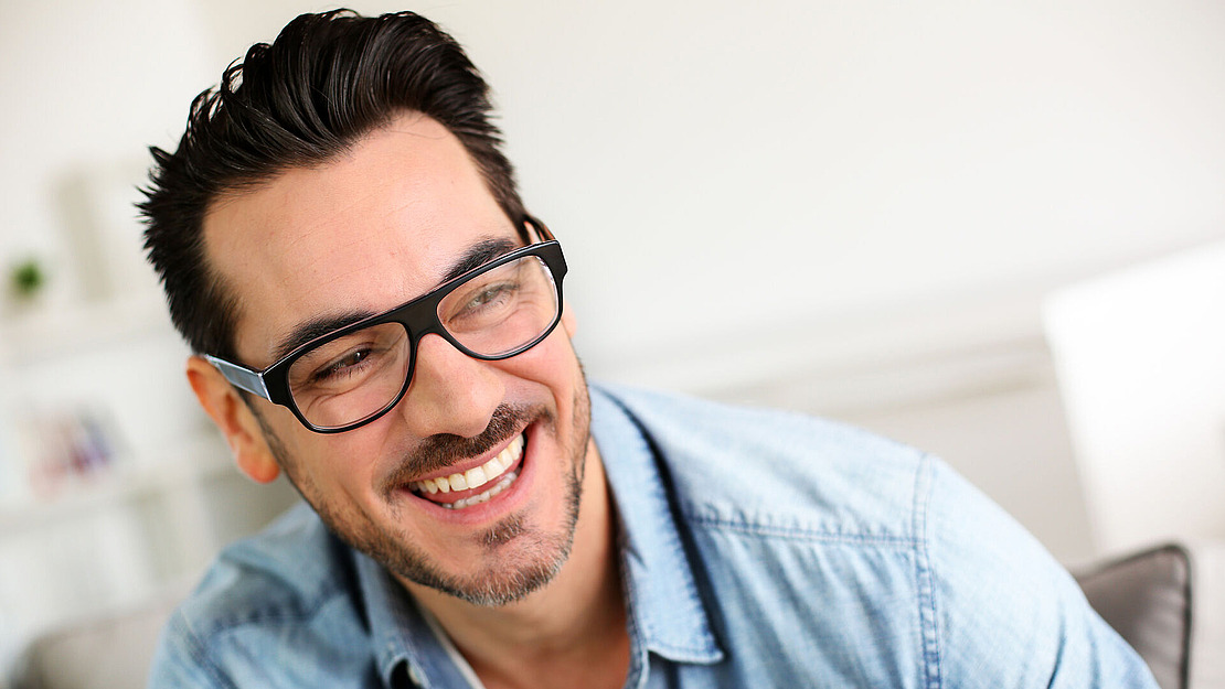 Ein lächelnder Mann mit kurzen dunklen Haaren, Brille und Gesichtsbehaarung, der ein hellblaues Jeanshemd trägt, sitzt in einem hell erleuchteten Raum und blickt zur Seite.