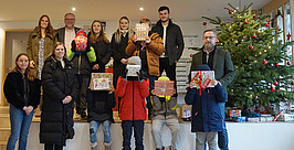Eine Gruppe von Erwachsenen und Kindern posiert in der Nähe eines geschmückten Weihnachtsbaums. Die Kinder vorn halten eingepackte Geschenke vor ihre Gesichter, während die Erwachsenen lächelnd hinter ihnen stehen.